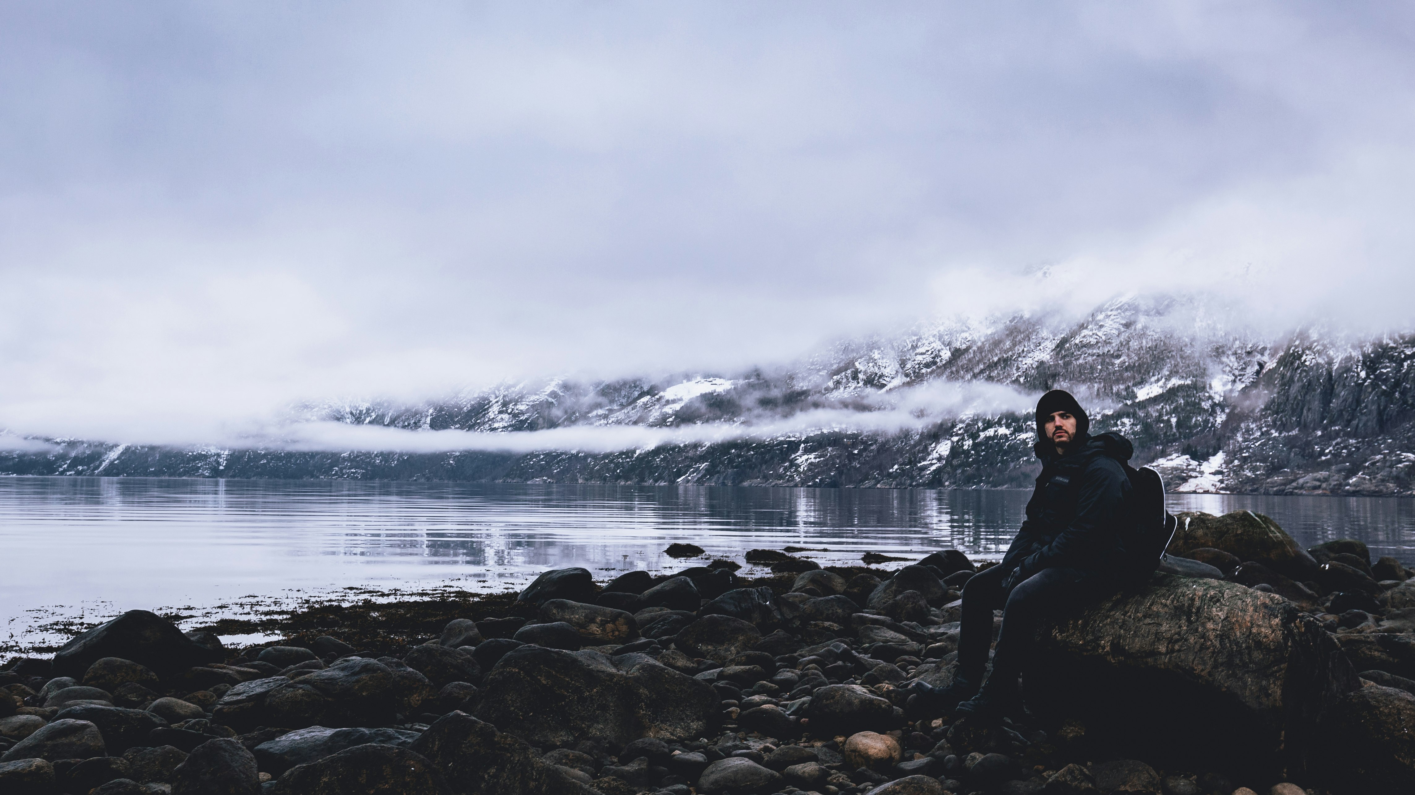 man sitting on rock overlooking calm body of water under white cloudy skies at daytime